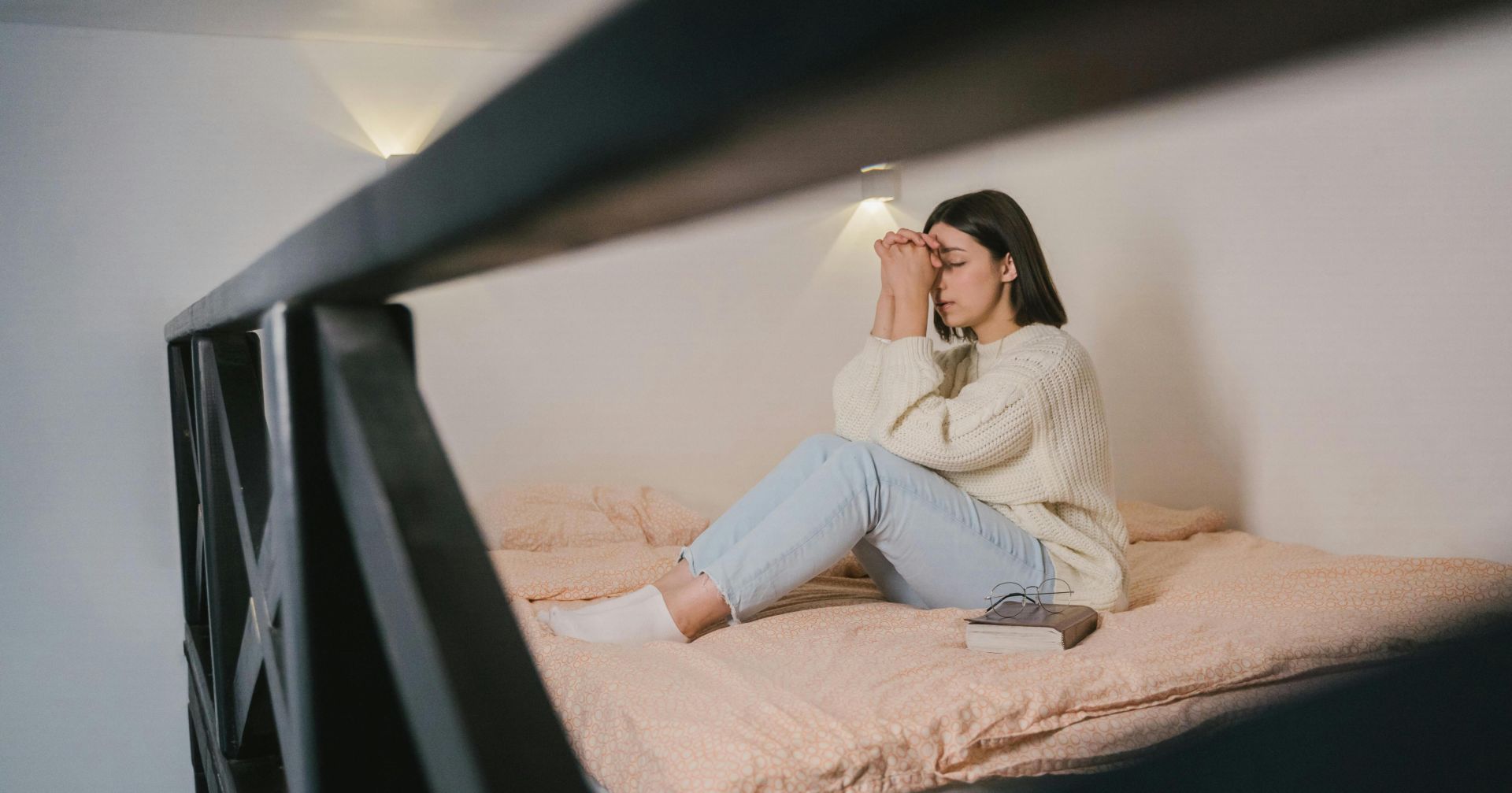 Woman in a cozy knitted sweater meditates peacefully on the bed with closed eyes and praying hands.
